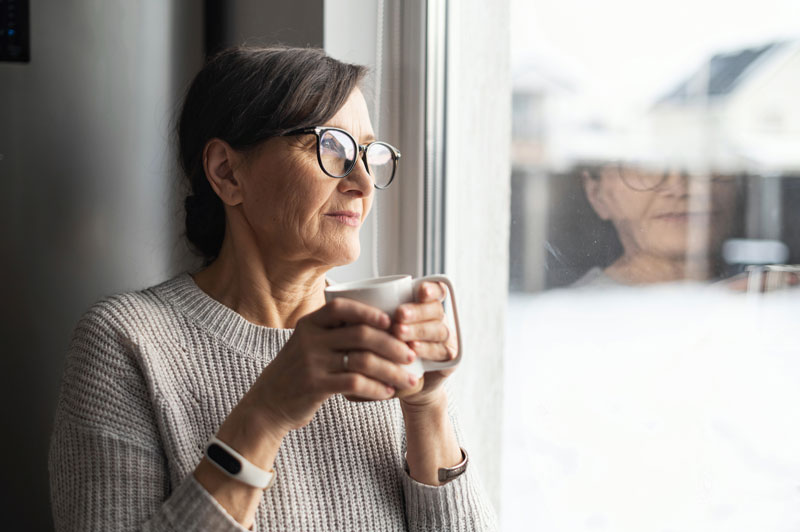 A woman holding a coffee mug stars earnestly out a window.