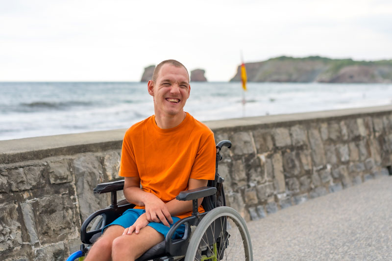 An Envigor NDIS client sits smiling in his wheelchair beside a stone wall at the beach