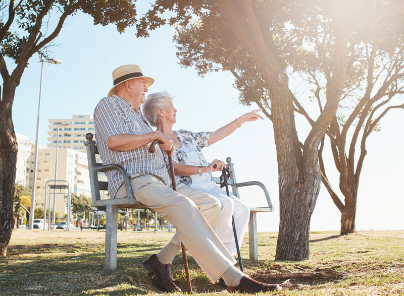 A happy, retired couple sit on a park bench looking out to sea.