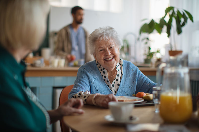 A smiling retirement village resident sits eating breakfast with her carer from a home care provider on the Sunshine Coast