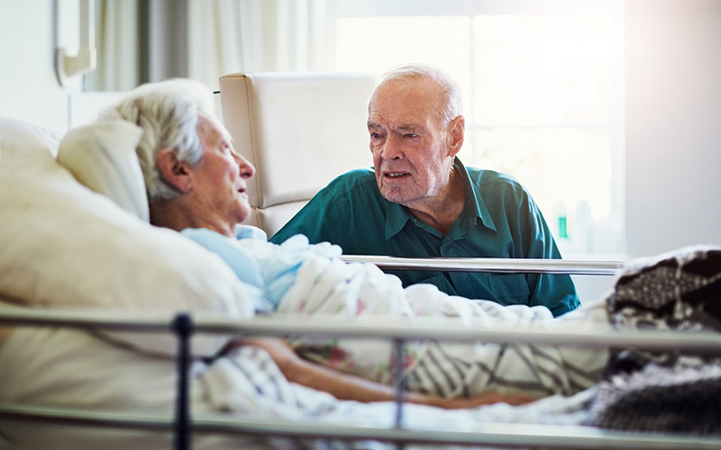 An elderly man sits with his wife at her bedside in hospital, as they prepare for her return home to recover.
